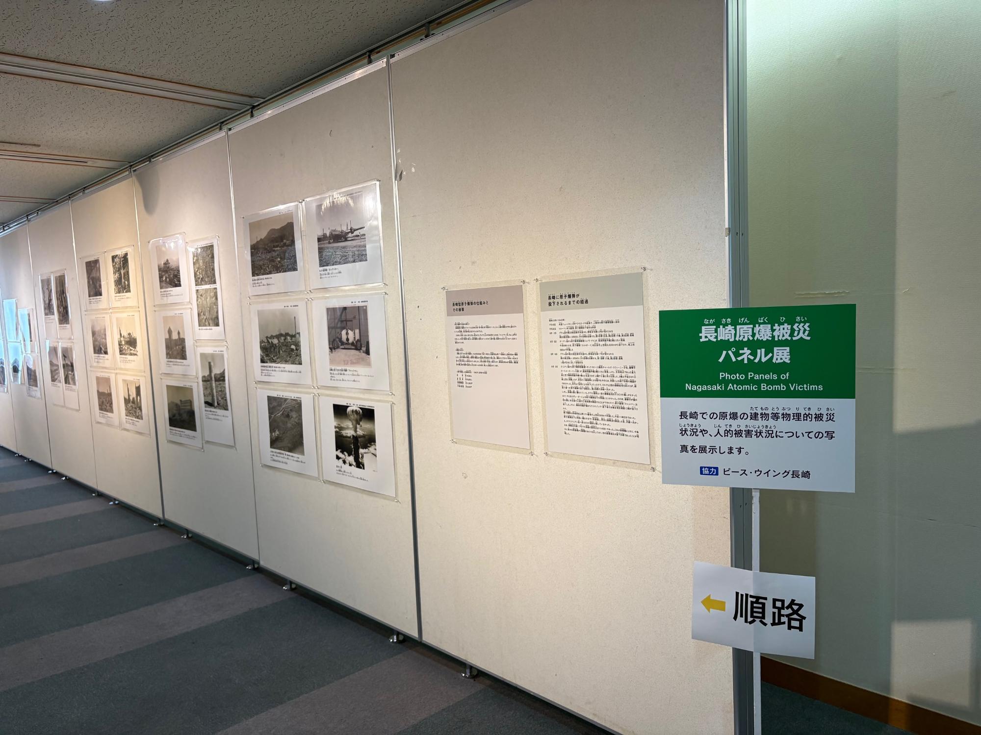 「長崎原爆被災パネル展」と書かれた緑色の案内看板の奥に、原爆投下直後の建物の被害状況や人々の被害を伝える多数のモノクロ写真がパーテーションに整然と掲示され、歴史の惨状を静かに伝える展示通路の風景の写真