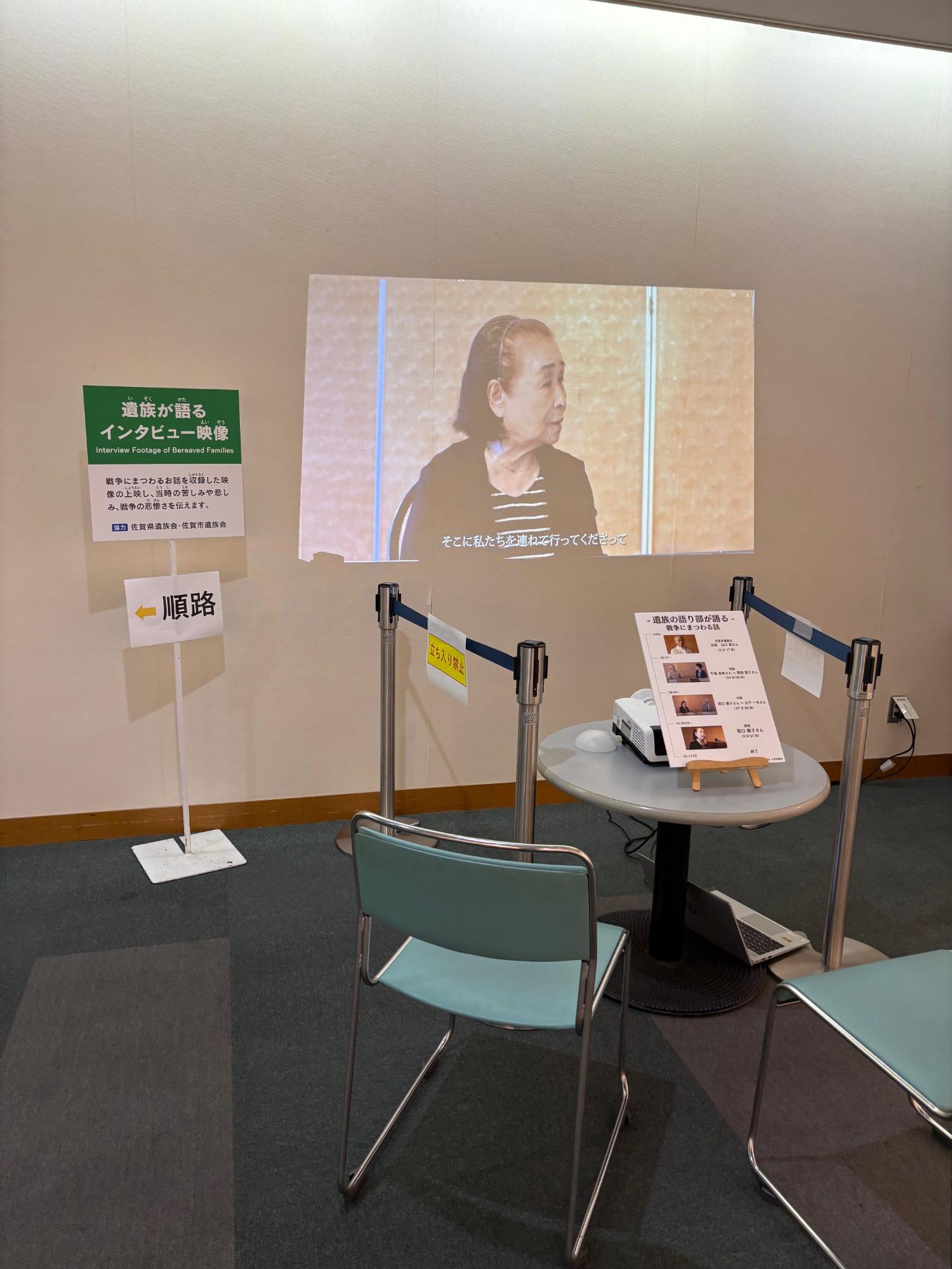 「遺族が語るインタビュー映像」という案内板の横で、壁面に投影された高齢女性の証言映像を椅子に座って視聴できるコーナーが設けられており、実体験を通して戦争の悲惨さを伝える展示空間の風景を捉えた写真