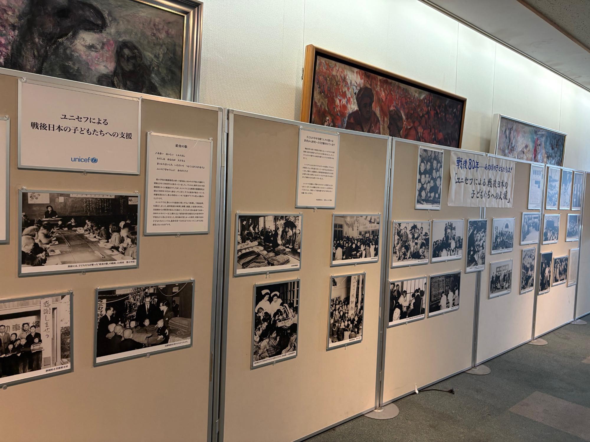 ユニセフによる戦後日本の子どもたちへの支援活動を伝える複数のモノクロ写真や解説パネルがパーテーションに並べられ、当時の給食風景や物資を受け取る様子を通して国際的な支援の歴史を紹介している展示風景の写真