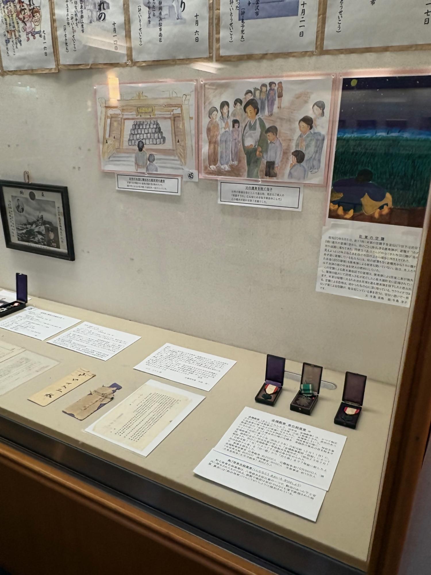 戦時中の様子を描いた絵画や解説文が壁面に掲げられ、手前の展示ケースには勲章や当時の文書、千人針などの遺品が丁寧に並べられており、過去の歴史と記憶を現代に伝える展示室の風景を斜めから捉えた写真
