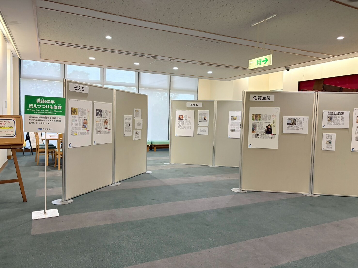 明るい展示室内に「戦後80年 伝えつづける使命」と題された緑色の案内看板が立ち、その奥には「伝える」「暮らし」「佐賀空襲」といったテーマごとに当時の新聞記事や解説パネルが整然と掲示されている風景の写真