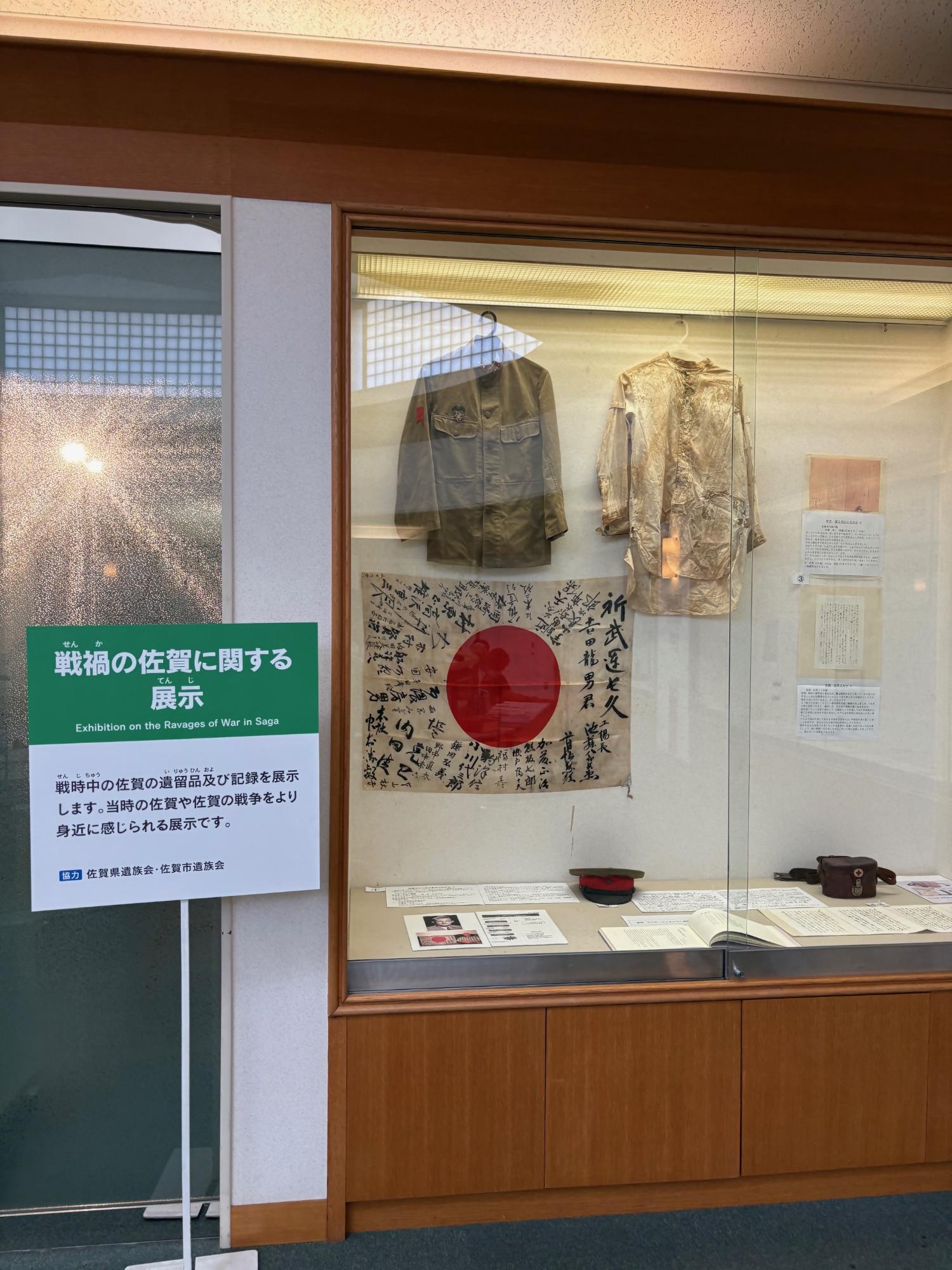 戦禍の佐賀に関する展示の案内パネルの横に、寄せ書きされた日の丸や軍服、当時の記録文書などの遺留品がガラスケース内に並べられ、戦時中の人々の暮らしや記憶を静かに伝えている展示風景の写真