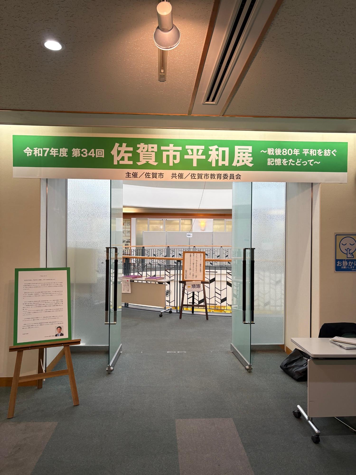 「令和7年度第34回佐賀市平和展」の緑色の看板が掲げられた会場の入り口で、ガラス扉の奥には展示スペースが広がり、手前には挨拶文が書かれたパネルや案内板が設置されている様子を捉えた写真