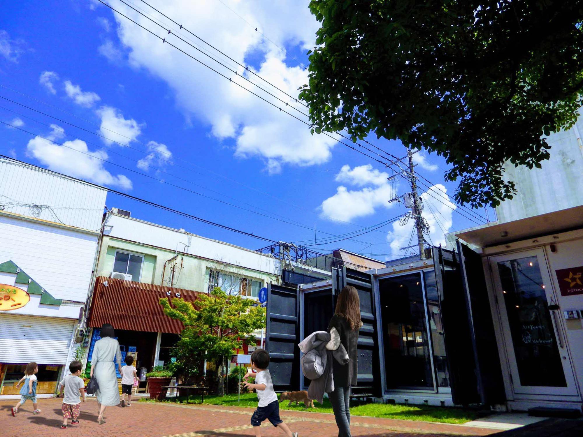 青空の下で商店が並ぶ広場を子どもたちが駆け回り大人が歩く中、木陰と建物が調和する街角の写真