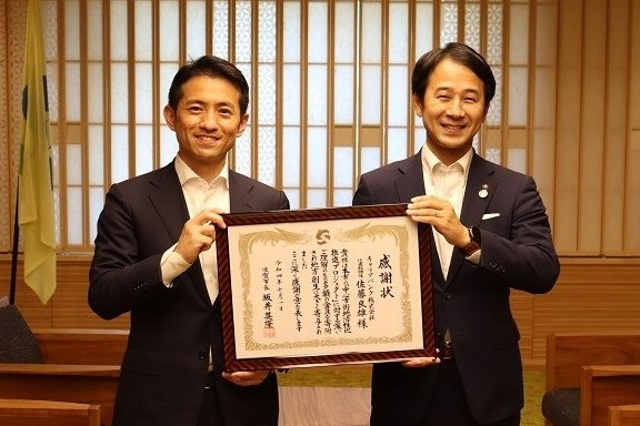 和風の木格子を背景に二人が額装された表彰状を共同で掲げて立つ授与場面の写真