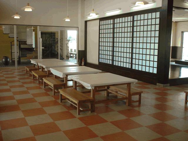 オレンジとベージュの市松模様の床が広がる室内で、白いテーブルクロスがかけられた木製の長机とベンチが並び、壁面には大きな障子戸やモダンな照明が設置された休憩スペースの写真