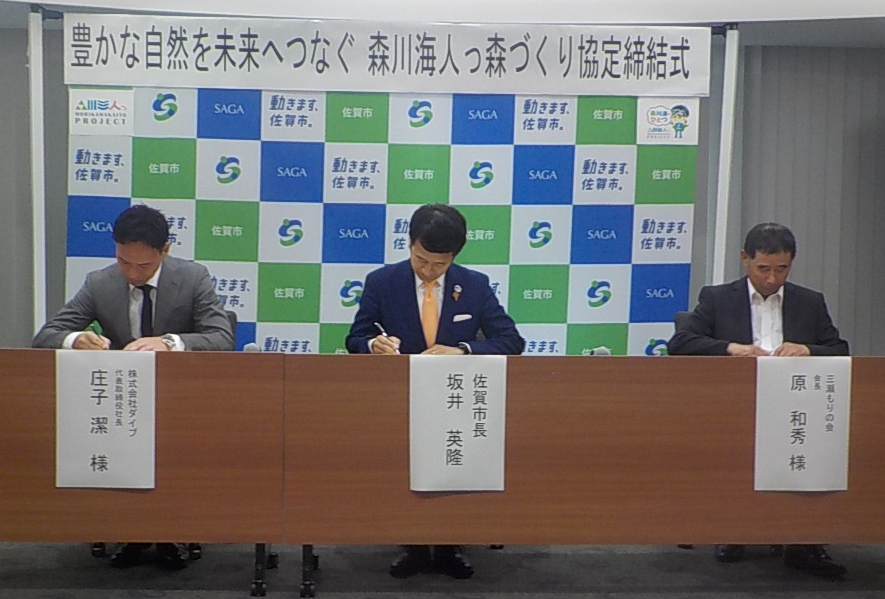 背後のパネルに「豊かな自然を未来へつなぐ森川海人っ森づくり協定締結式」と書かれ、着席した3人が机に並んで書類へ署名する様子の写真