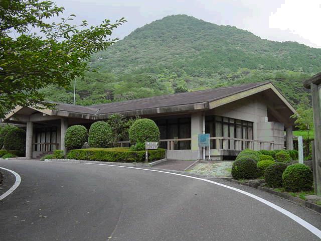 緩やかなカーブを描くアスファルトの道路沿いに建つ、落ち着いた色調で平屋建てのモダンな建物と、背後にそびえる緑豊かな深い山々が一体となって静かな自然環境を醸し出しているセンターハウスの写真