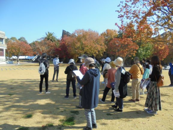 背後に色鮮やかに紅葉した木々が広がる穏やかな秋の木漏れ日が差し込む芝生の上で、資料を手にしたガイドの説明を十数人の参加者が等間隔で囲むように聞いている様子の写真
