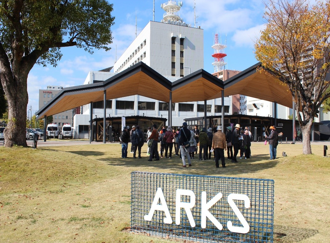 青空が広がる芝生広場で、手前に「ARKS」のロゴ看板が立つ中、特徴的なギザギザ屋根の大きな東屋の下に集まった防寒着姿の参加者たちが、周囲の建物や景観についてガイドから説明を受けている写真