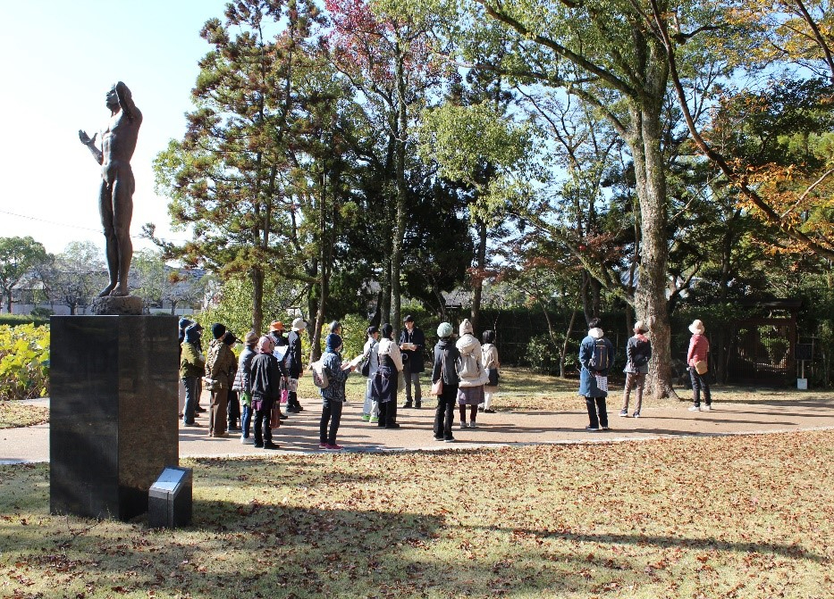 明るい日差しが注ぐ秋の公園で、台座に立つ男性の裸体彫像を左手に見ながら、厚手のコートや帽子を身にまとった大勢の参加者たちが、木々に囲まれた散策路でガイドの説明に耳を傾けている写真