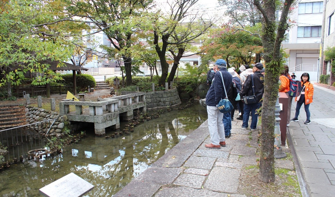 紅葉が始まった木々に囲まれた静かな水辺で、石造りの護岸や歴史を感じさせる構造物を眺めながら、オレンジ色のジャンパーを着たスタッフと共に歩道に並んで地域の歴史を学ぶ、ウォーキングイベントの一場面を捉えた写真