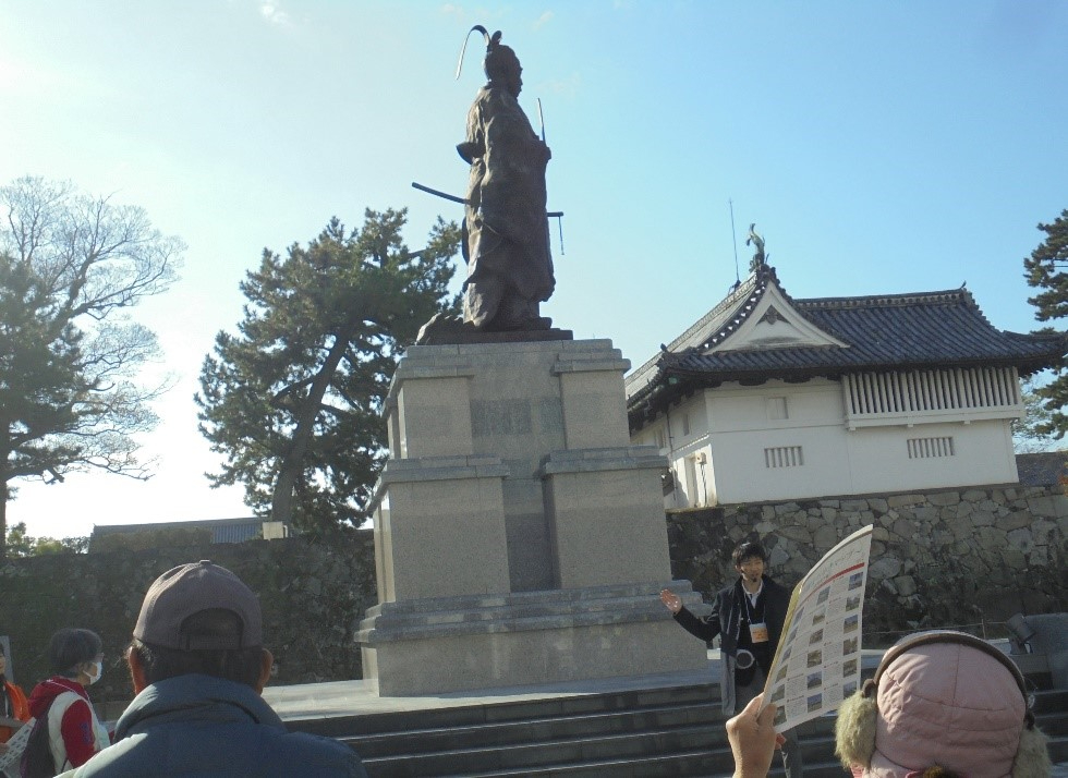 青空を背に高い石垣の上に立つ鍋島直正公銅像を見上げながら、ガイドによる解説を熱心に聞き入る観光客たちの様子と、その奥に歴史を感じさせる白壁の日本建築が佇む佐賀城本丸歴史館周辺の風景写真