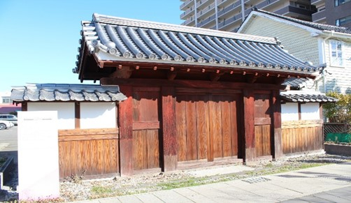 伝統的な瓦屋根を載せた木製の門が陽光に照らされ、周囲の現代的建物と対比を成す旧嬉野家の武家屋敷門の写真