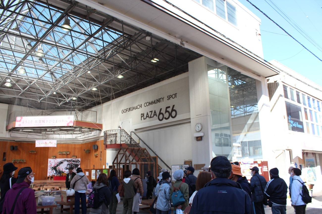 屋根付き広場の中央に人々が集まり、PLAZA656の看板や木製ステージが見える写真