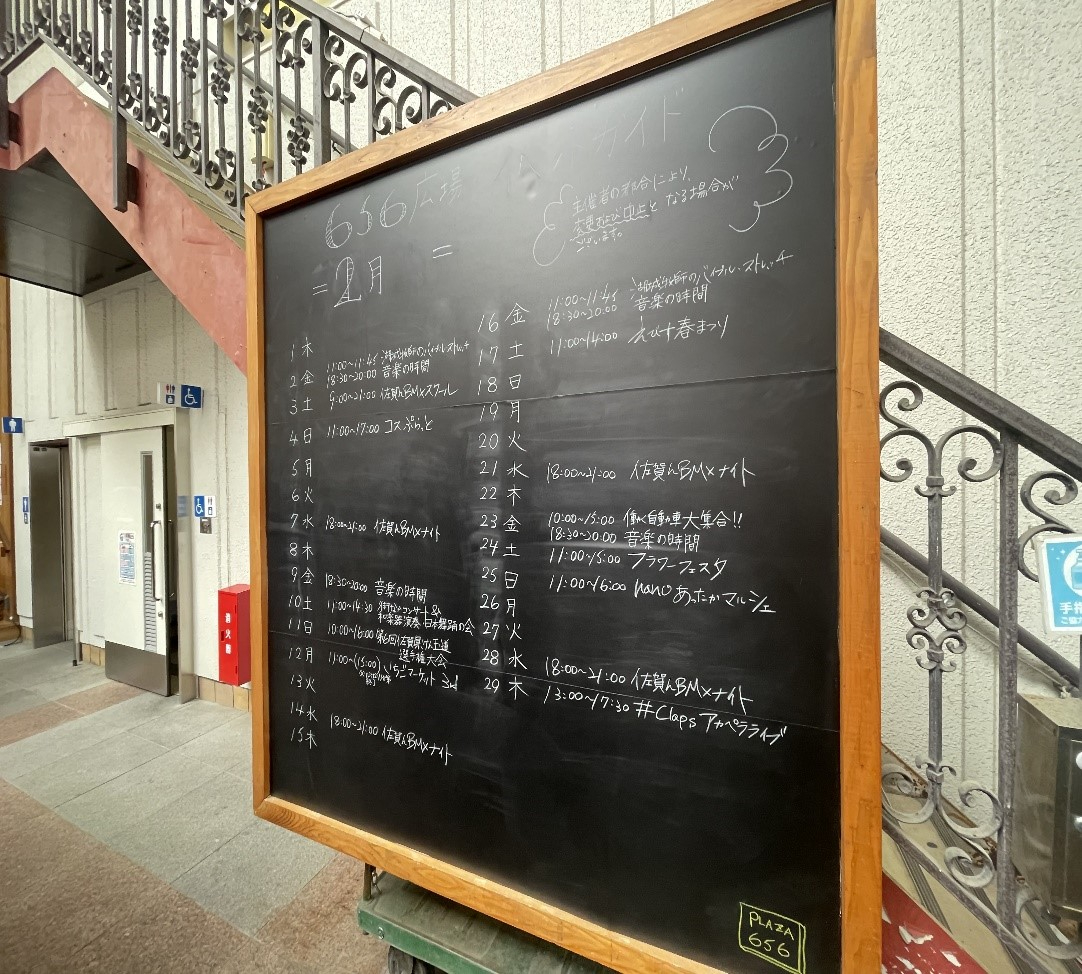 黒板に手書きのイベント日程が月ごとに細かく記され、周囲に階段やエレベーターが見える屋内掲示の写真