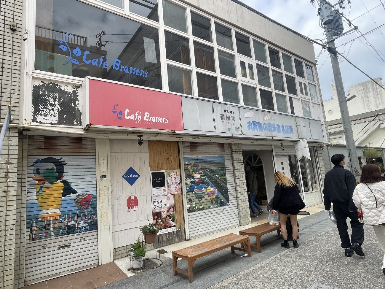 古い商店街の一角で、カフェの看板やシャッターの彩色画が並ぶ二階建ての建物前を数人の通行人が歩いている写真