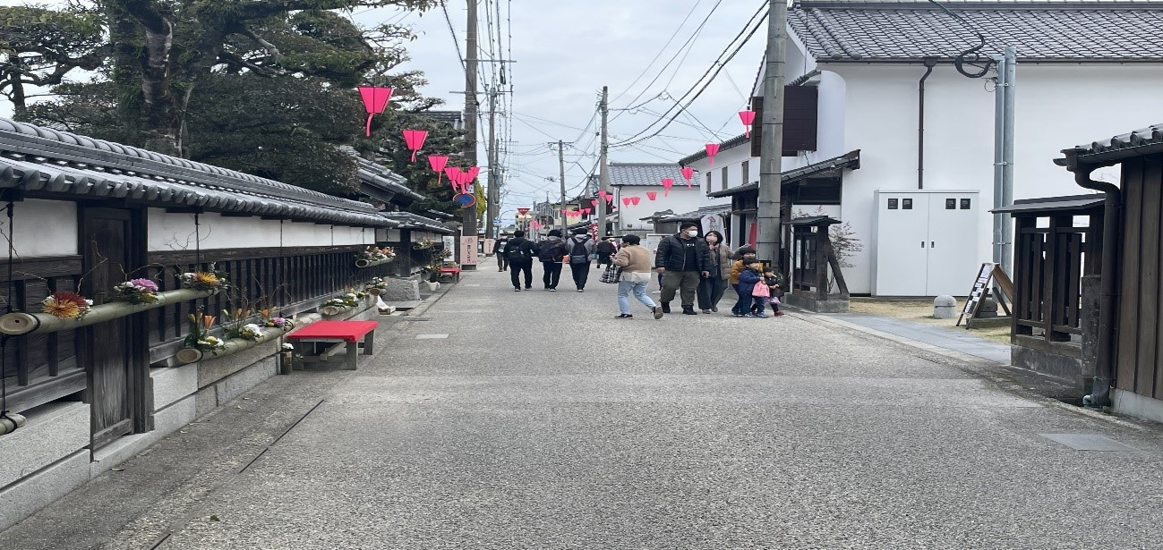石造りの道沿いに古い町家が並び、花飾りや赤い提灯風の旗の下を人々が歩く柳町通りの写真