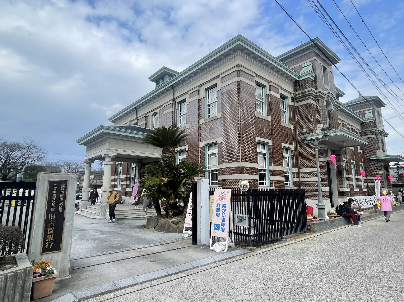 赤レンガの重厚な洋風建築が青空の下に佇み来訪者が旧古賀銀行入口周辺を行き交う様子と歴史的雰囲気が漂う情景を映した写真