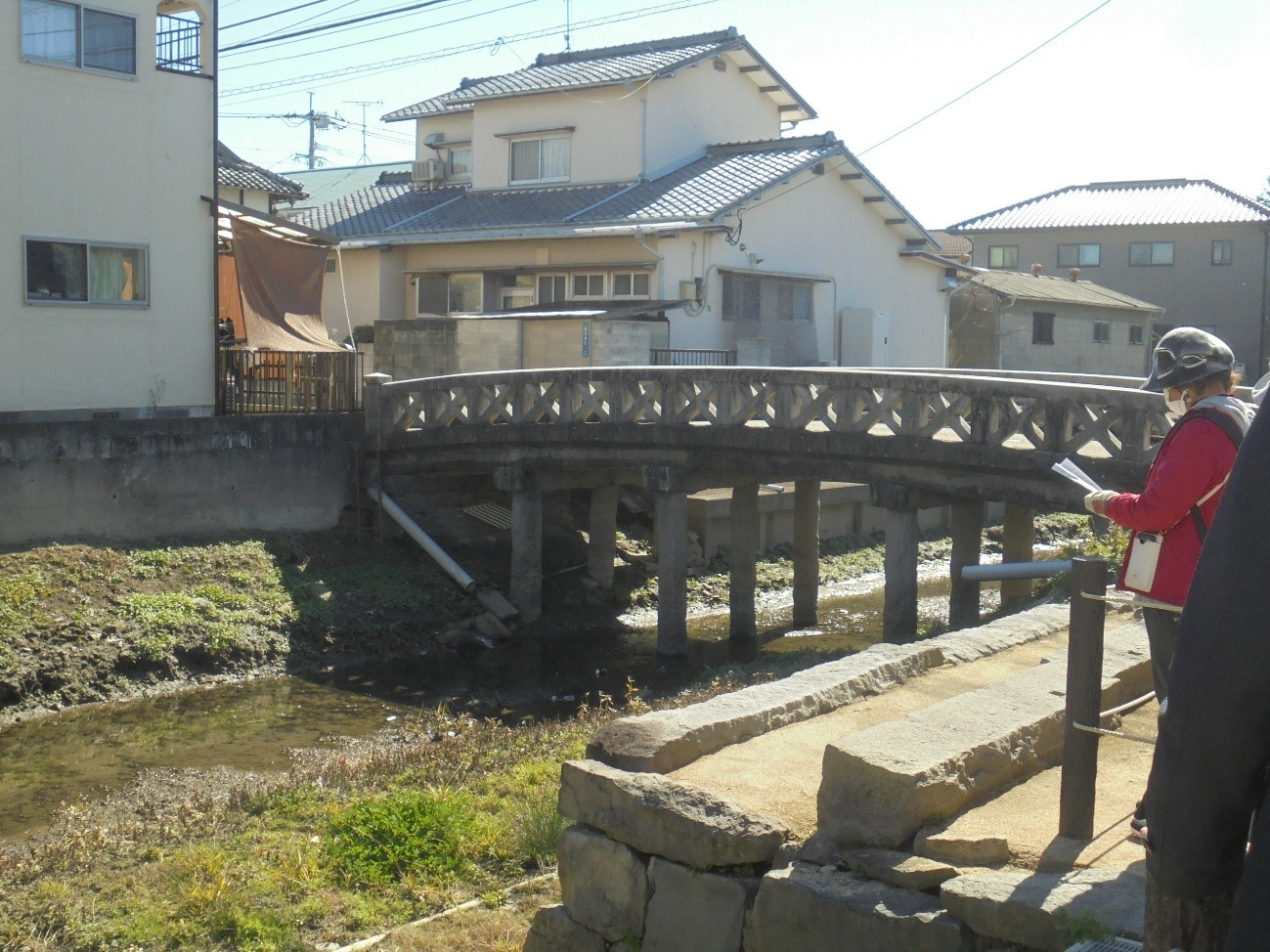 用水路に架かる木製風の古い橋と周囲の住宅を背景に見学者が資料を手に佇む写真