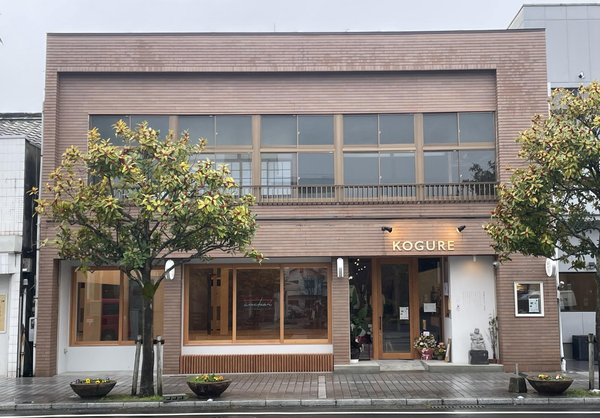 曇り空の下、歩道沿いに建つ茶色のレンガ風外壁の2階建て店舗ビルがあり、木枠の大きな窓と「KOGURE」のロゴが掲げられ、左右には緑の葉と赤い実をつけた2本の植栽が配置されている株式会社RELI.STYLEの建物外観の写真