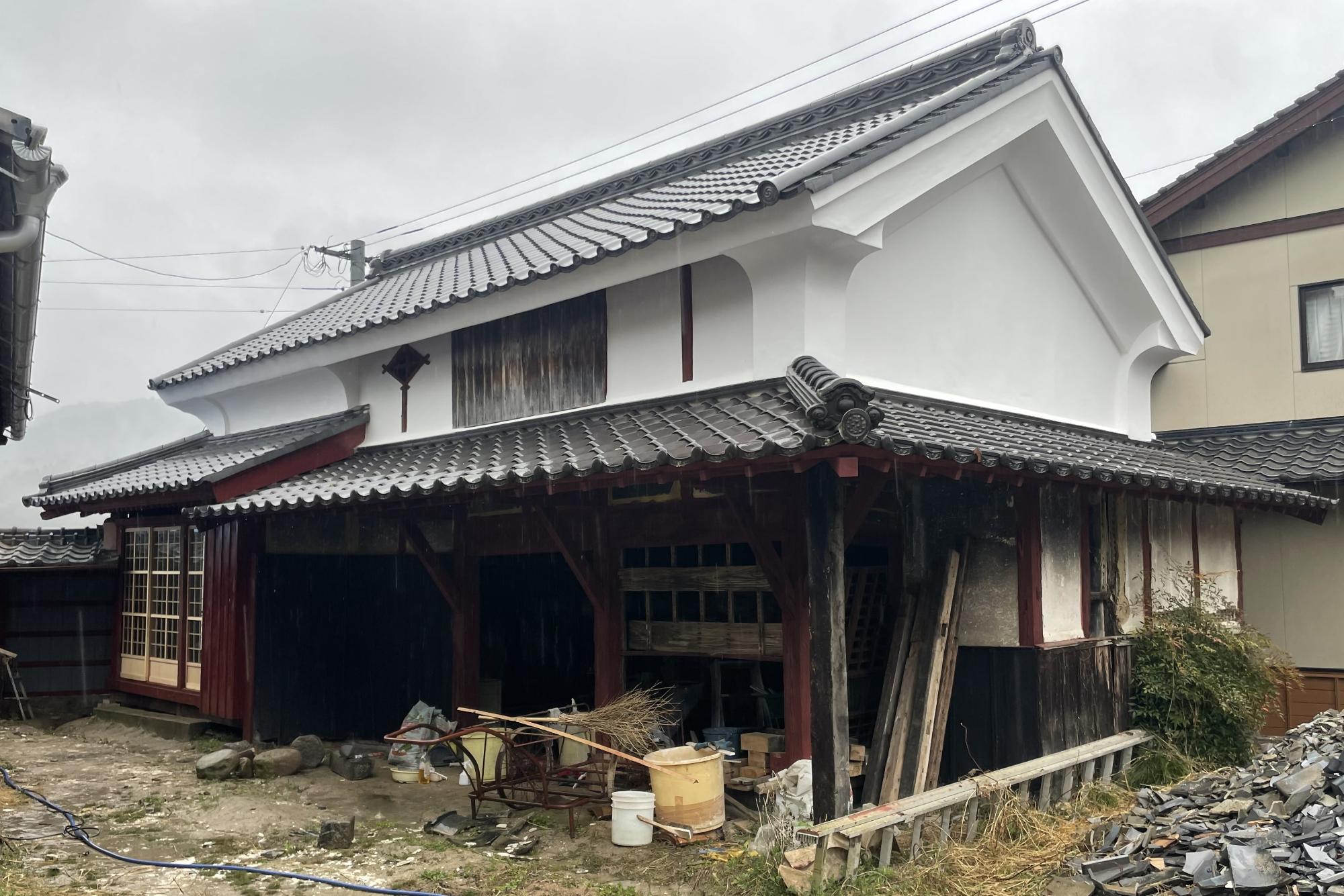 瓦屋根と白壁の蔵のような古い木造建築があり、軒下には資材や道具が置かれ、手前の地面には瓦の破片が積み重なっている、曇り空の下で撮影された歴史を感じさせる旧松野家住宅の建物外観の写真