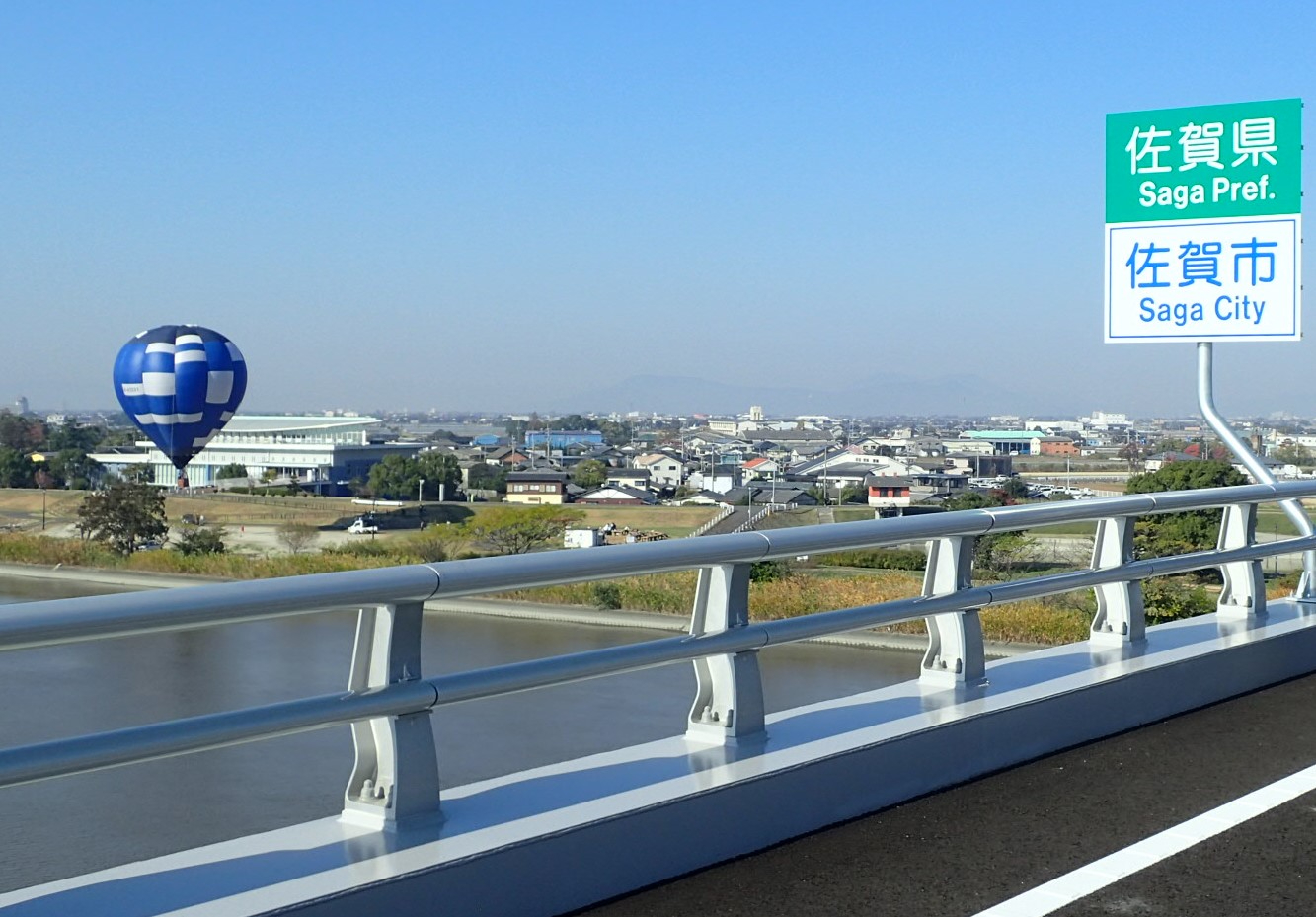 青空の下、川沿いの道路から佐賀県と佐賀市の標識越しに青白の熱気球と市街地を望む写真