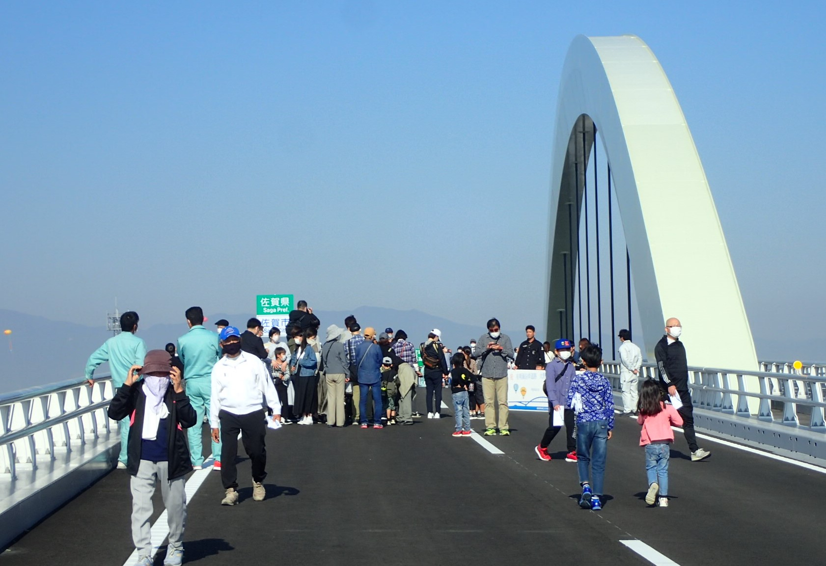 青空の下、新しい大きなアーチ橋の車道を多くの人々が行き交いながら開通前の雰囲気を楽しむ様子の姿の写真