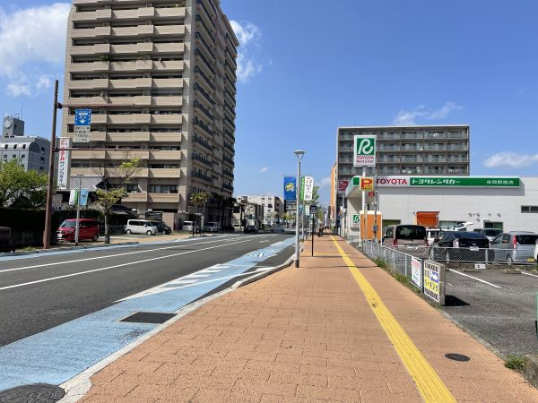 青空の下、左側に高層マンション、右側にトヨタレンタカーの店舗が並ぶ平坦な道路において、手前から奥へと続く茶色のタイル舗装された広い歩道と、青いペイントが施された自転車優先レーンがある車道の風景を捉えた写真