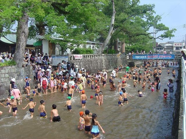 青空の下、緑豊かな樹木に囲まれた浅い川や水辺で、大勢の子どもや大人が水遊びを楽しみ、周囲に見物客や横断幕が見える多布施川水遊び場の写真