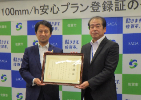 佐賀市のロゴが並ぶ背景パネルの前で坂井市長と野村真一氏が額入りの登録証を丁寧に掲げて並んで立つ記念撮影の写真