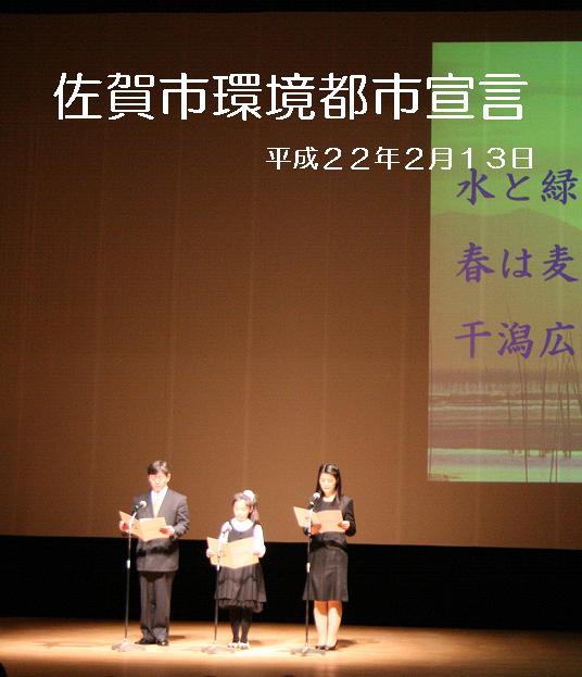 ステージ上で3名の発表者が佐賀市環境都市宣言平成22年2月13日と水と緑春は麦秋干潟広がるのスクリーン映像を背景に原稿を手に宣言を読み上げている式典の写真
