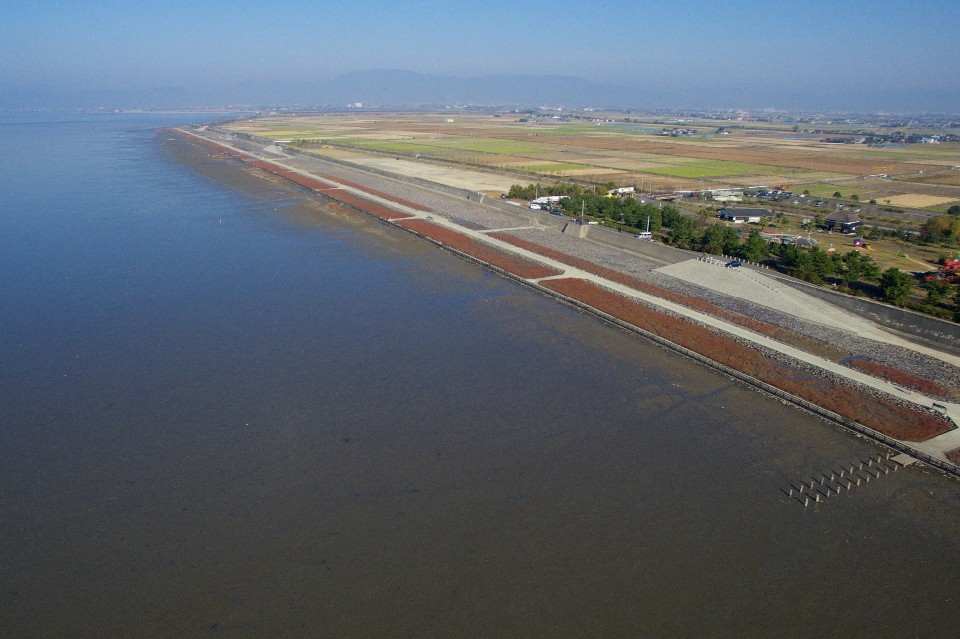 穏やかな水面が広がる海岸線に沿って、赤茶色の植物が群生する長い堤防と整備された遊歩道が続き、その奥には広大な田畑や街並み、そして霞んだ山々が地平線まで広がる風景を上空から見渡した写真