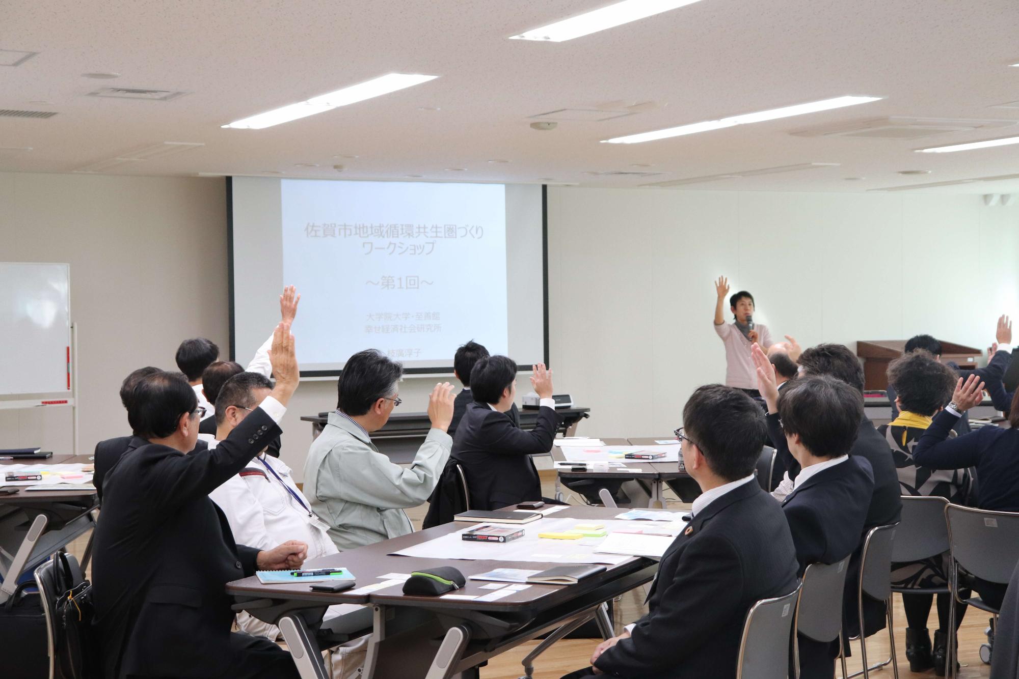 明るい会議室で、スーツ姿の参加者たちがテーブルに座り、正面のスクリーンに佐賀市地域循環共生圏づくりワークショップ第1回と投影され、右側に立つピンクシャツの講師に向かって多数の参加者が挙手をしている写真