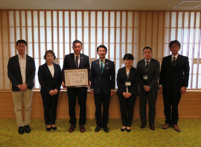 和室を背景にスーツ姿の7名が一列に並び、中央の田中鉄工の代表の方が表彰状を持って記念撮影に臨んでいる感謝状贈呈式の写真
