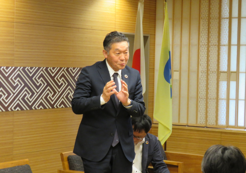 和室の会議室で日本国旗と自治体旗を背景に、スーツ姿の田中鉄工の代表の方が立って手を合わせるジェスチャーで話をしている様子の写真