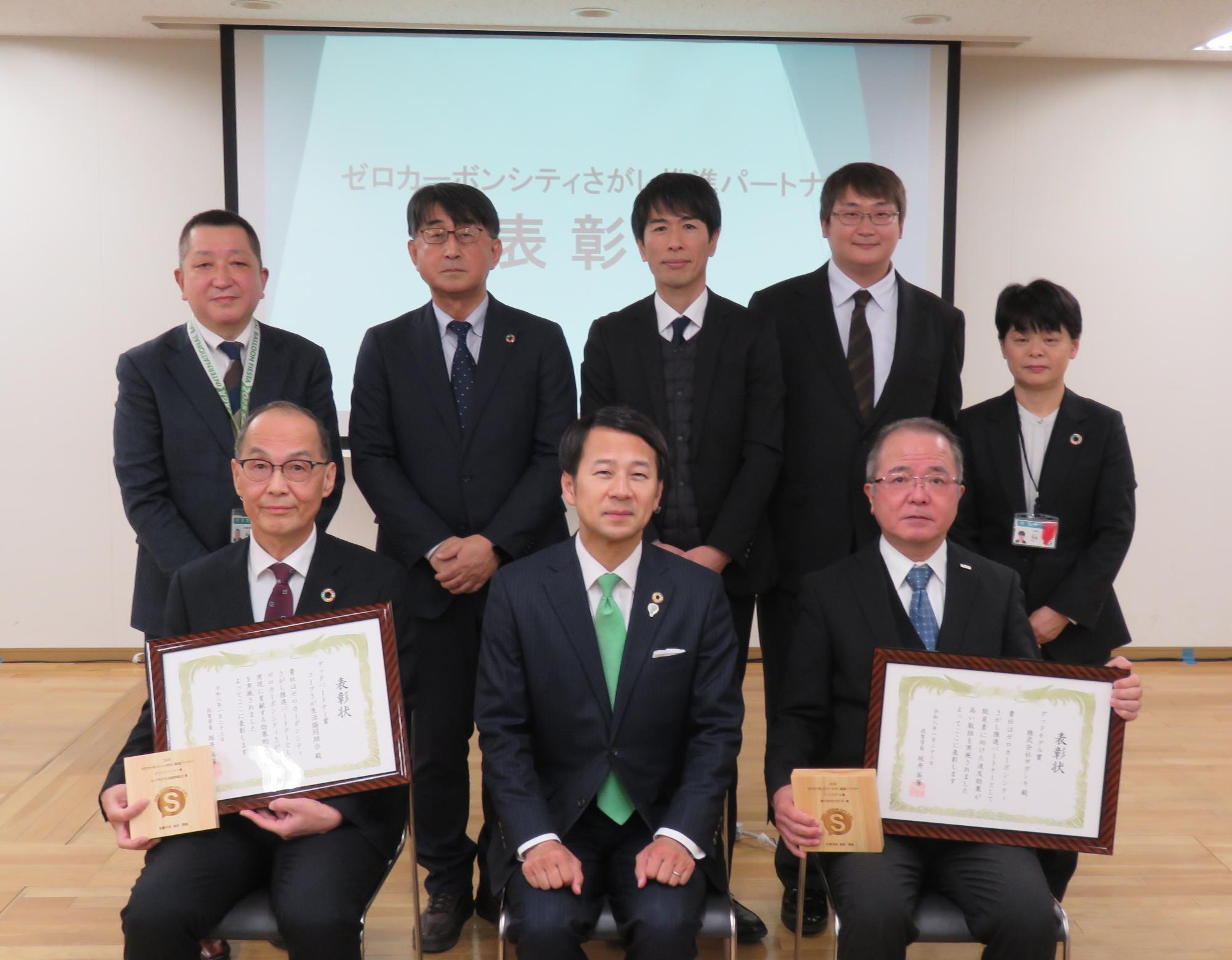 「ゼロカーボンシティさがし推進パートナー表彰」式典で、スーツ姿の男女8人が椅子に座る3人(中央が佐賀市長)と後ろに立つ5人とで整列し、前列両端の2人がそれぞれ表彰状とマークの入った木製の盾を持っている記念写真