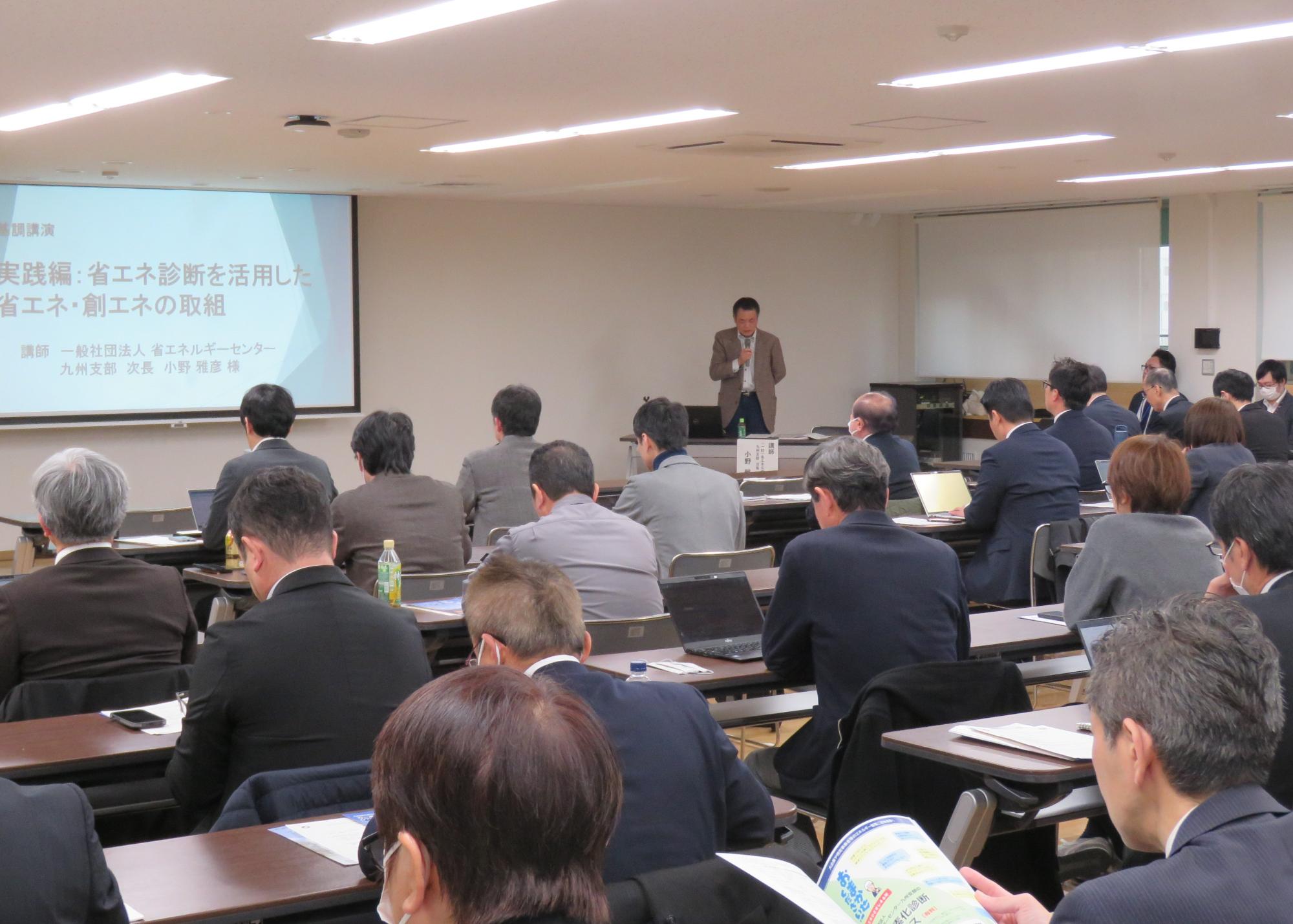 「省エネ診断を活用した省エネ・創エネの取組」と題されたスライドを背に講師がマイクで話し、多くの参加者が熱心にメモを取ったりパソコンを操作したりしながら聴講している講演会の様子を捉えた写真
