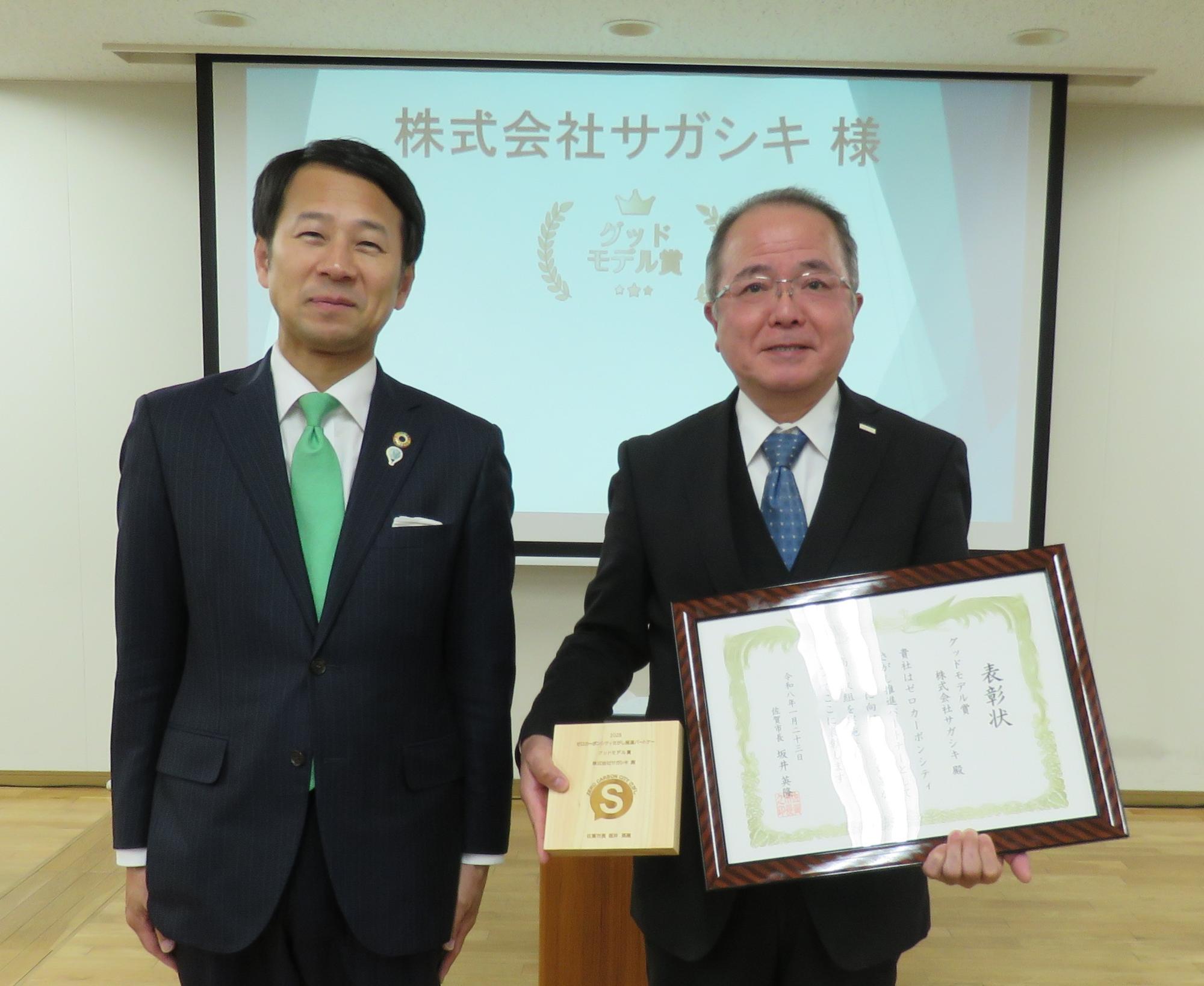 スクリーンに株式会社サガシキとグッドモデル賞のロゴが投影された室内で、市長と株式会社サガシキの代表の方が額装された表彰状と木製の盾を持ち記念撮影している写真