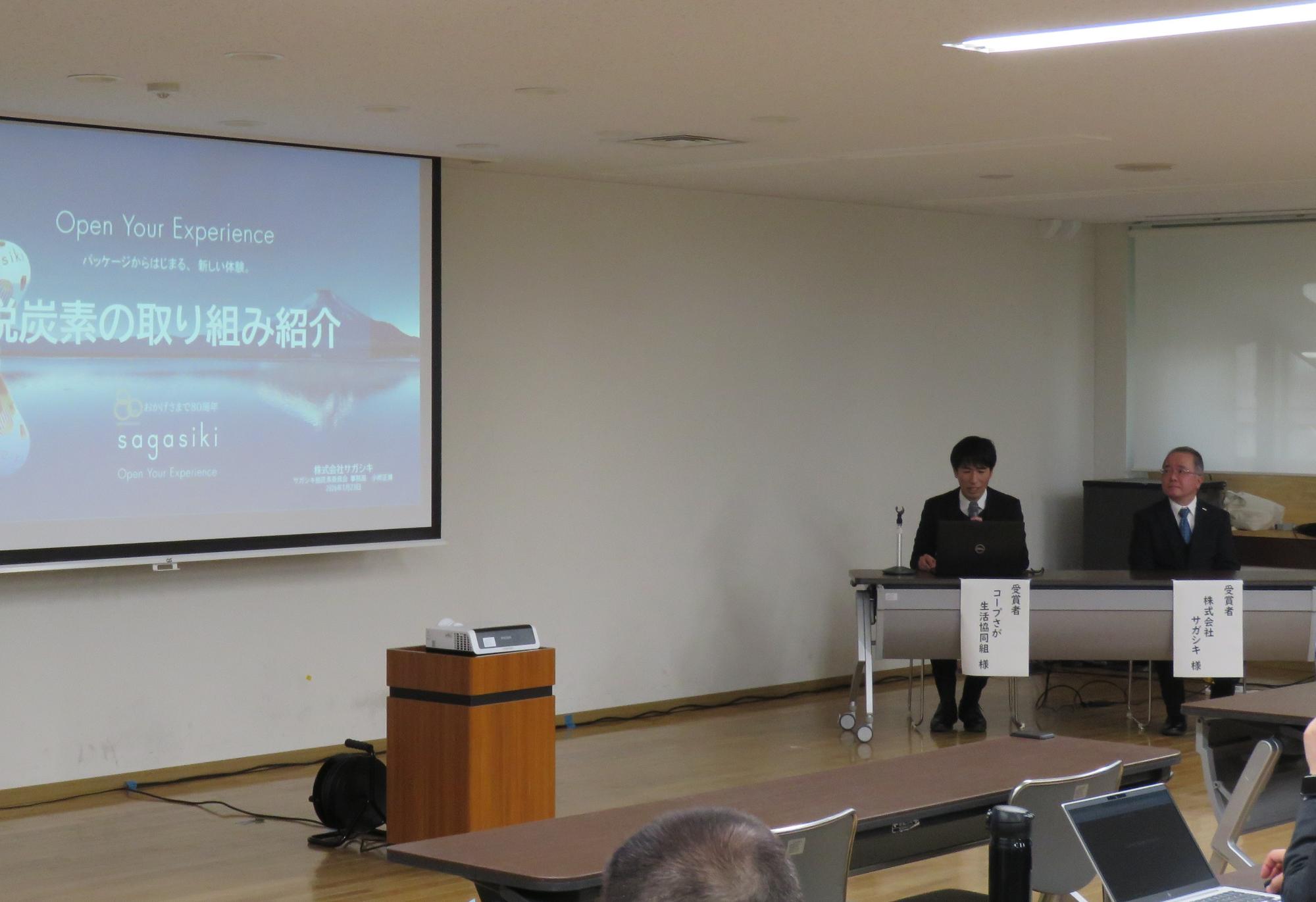 「脱炭素の取り組み紹介」と書かれたスライドが投影されたスクリーンの横で、スーツ姿の男性二名が机に座り、ノートパソコンを操作しながら取り組みを紹介し進行している会議室の風景を捉えた写真