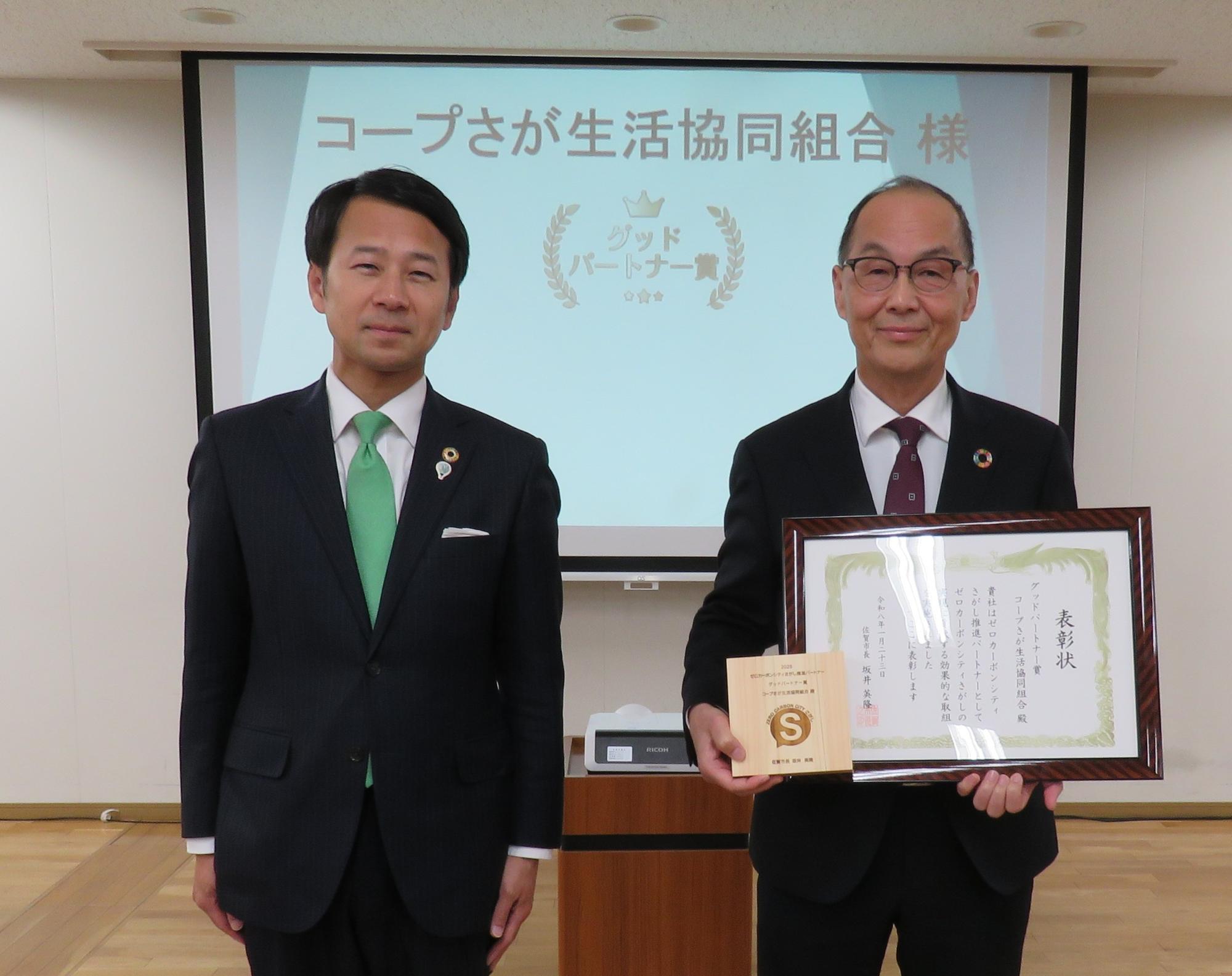 「グッドパートナー表彰」と書かれたスクリーンを背景に、市長とコープさが生活協同組合の代表の方が表彰状と「S」マークの入った木製の盾を持って正面を向いて記念撮影している写真