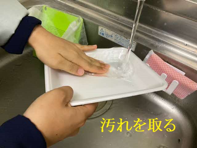 キッチンのシンクで蛇口から流れる水を使用し、リサイクルに出すための白い食品トレーを素手で丁寧に水洗いして、表面に付着した残菜などの汚れを落としている様子の写真