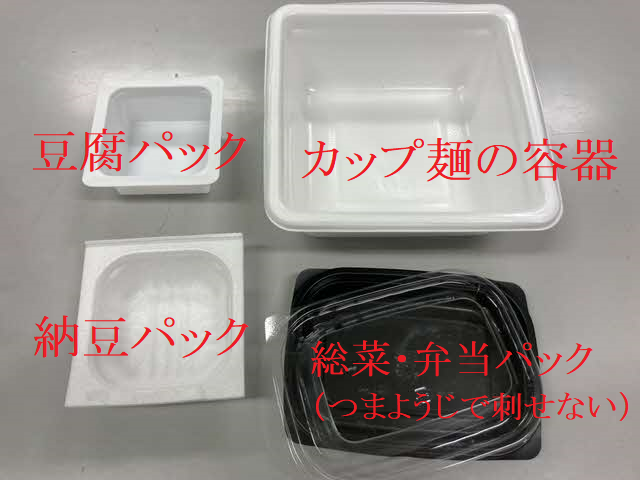 平らな作業台の上に、豆腐パック、納豆パック、カップ麺の容器、そして「つまようじで刺せない」と赤字で注釈が添えられた透明な蓋付きの黒い総菜・弁当パックが並べられた、素材の違いを比較する写真