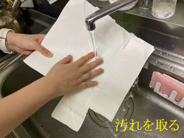 キッチンのシンクで蛇口から流れる水を使用し、リサイクル回収に出すために切り開いた白い牛乳パックの内側を素手で丁寧に水洗いして、付着した汚れをしっかりと落としている様子の写真