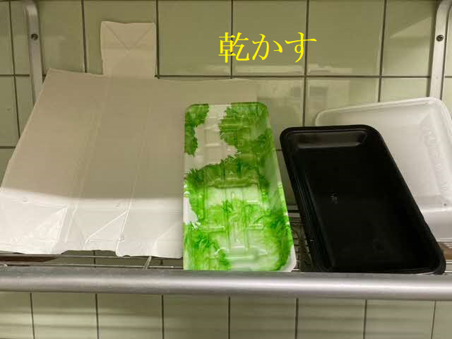 水洗いした後の白い牛乳パックや、緑色の柄入り、黒色、白色の様々な食品トレーが、キッチンの水切りラックに立てかけられ、上部に黄色い文字で「乾かす」と書かれた写真