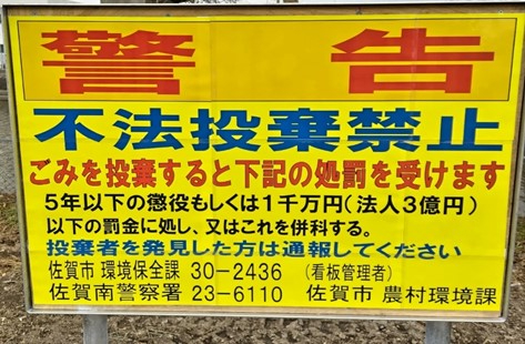 黄色の背景に赤字で大きく「警告」、青字で「不法投棄禁止」と記され、違反時の罰則内容や発見時の通報のお願い、佐賀市環境保全課や警察署の連絡先が詳細に記載された、不法投棄を強く抑止するための警告看板の写真