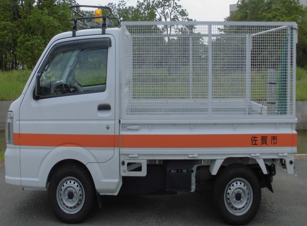 白い車体にオレンジ色のラインが入り、後部には「佐賀市」の文字が刻まれ、荷台部分に不法投棄物の回収用と思われる白い金属製のフェンスが取り付けられた、作業用の軽トラックを真横から捉えた写真