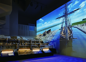 薄暗い展示室内に、三重津海軍所跡の木組遺構の実物展示があり、その右側の壁面にはかつてのドックや帆船の様子を鮮やかな色彩で描いた巨大なシアター映像が投影されている、歴史と最新技術を融合させた博物館の展示風景の写真
