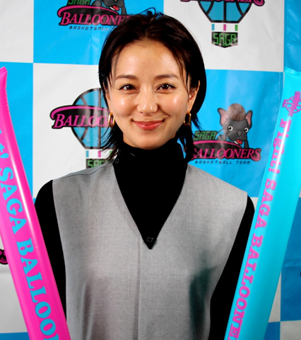 水色と白の市松模様に「SAGA BALLOONERS」のロゴが並ぶ背景の前で、黒のタートルネックにグレーのベストを着た中越典子さんが、ピンクと水色の応援用バルーンスティックを手に持ち微笑むポートレート写真