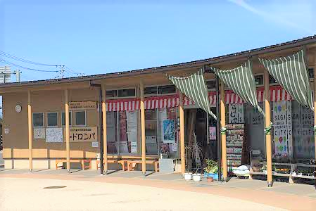 青空の下、ベージュの外壁に「橋の駅 ドロンパ」の看板が掲げられた平屋の建物があり、軒先には緑と白のストライプ柄の日除けが並び、ガラス窓に赤と白の装飾やポスターが貼られた賑やかな店舗外観の写真(橋の駅 ドロンパのサイトへリンク)